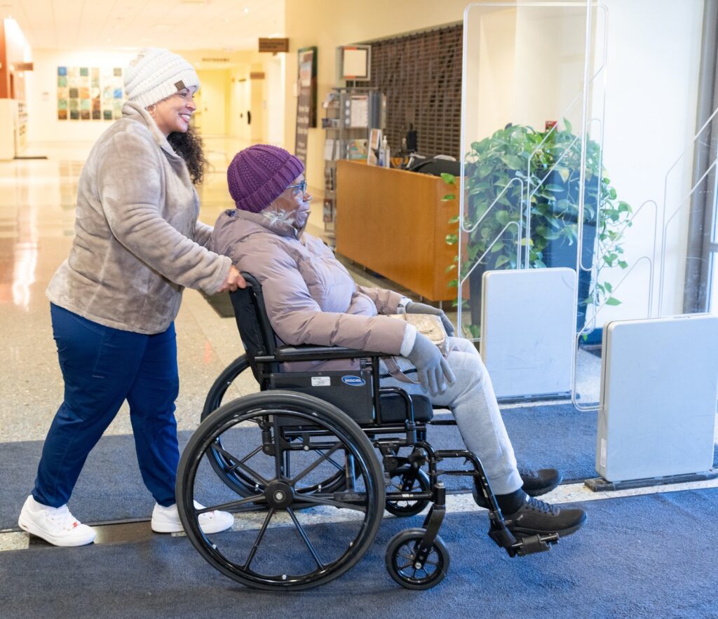A Care Partner walking with a client using a wheelchair in a bright lobby